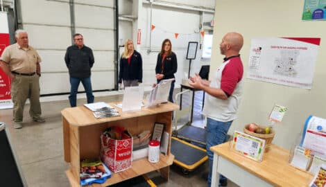 A stocked pantry cart of donated food beside a table of produce as food bank staff and visitors talk in a warehouse