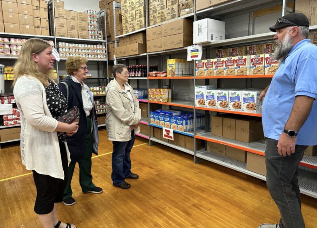 Three visitors listen to a guide in a blue polo shirt beside shelves stocked with cereal boxes and canned goods inside the New Love Center food pantry