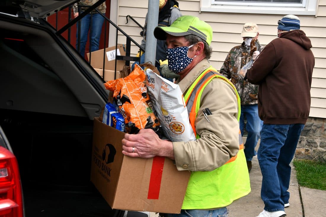 A masked volunteer in a safety vest loads a Central Pennsylvania Food Bank box of food into a car trunk during the Good Neighbor Center's drive-up distribution in Renovo, PA