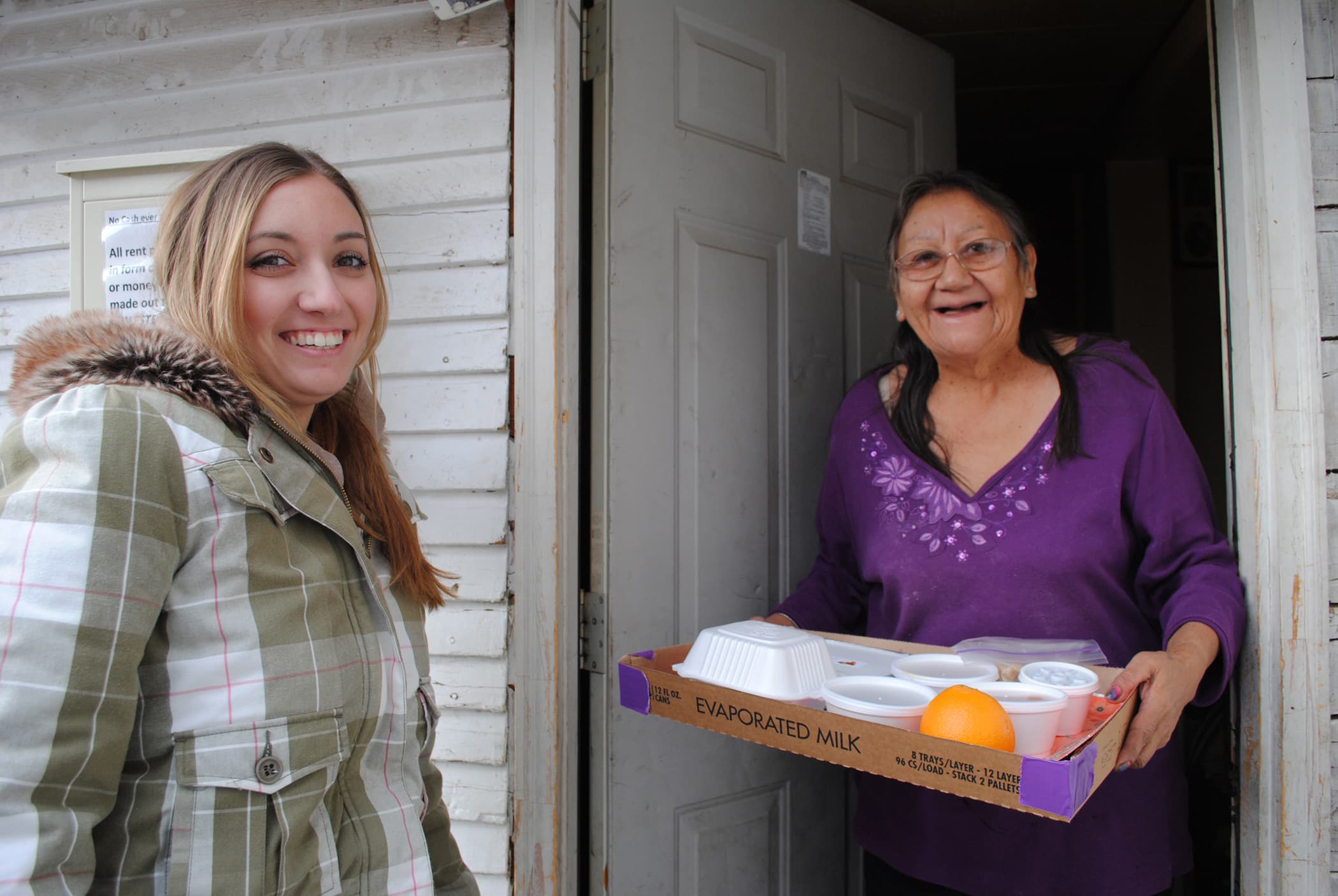 An older woman smiles as a uniformed volunteer hands her a Meals on Wheels meal at her front door