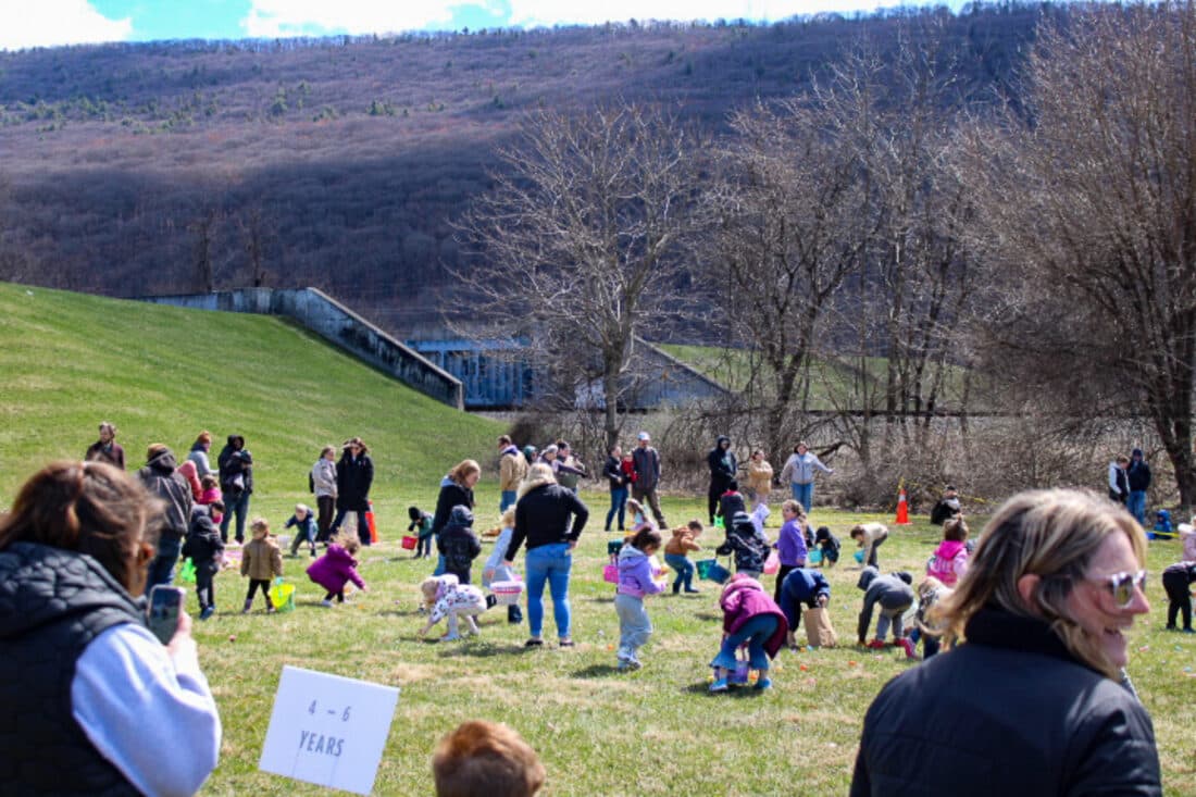 Families and young children gather Easter eggs on the grass at the Castanea Fire Company picnic grounds during the company's first egg hunt, with a wooded ridge rising behind the field